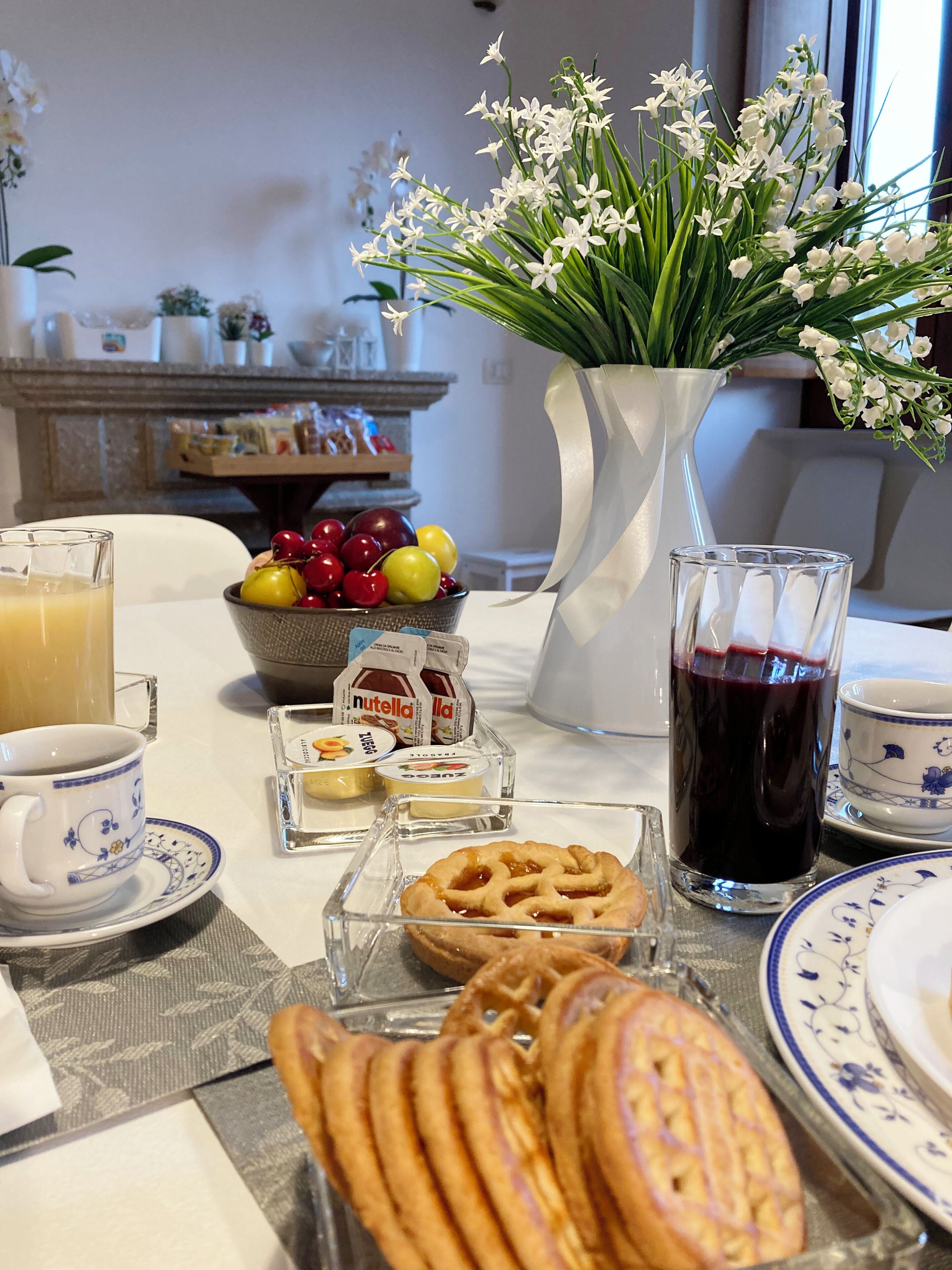 A breakfast setting with a table, a vase of flowers, a bowl of fruit, a glass of juice, a cup of coffee, a plate with pastries, and a jug of milk.