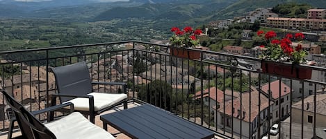 A balcony with a table and chairs, overlooking a mountainous landscape with buildings.