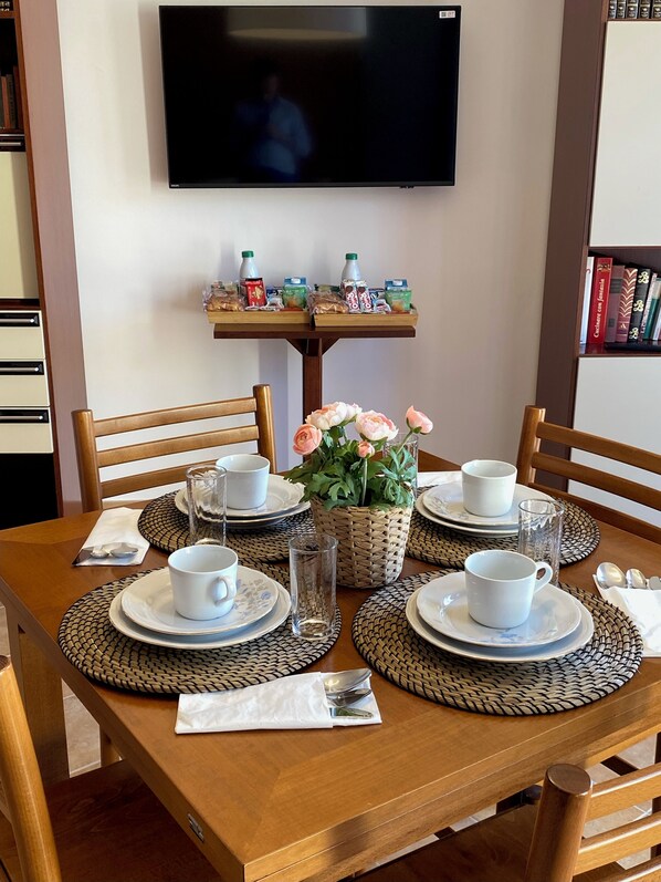 Une table à manger est dressée avec des tasses, des soucoupes et des assiettes blanches, entourée de sets de table en fibres tressées. Un panier rempli de fleurs est placé au centre.