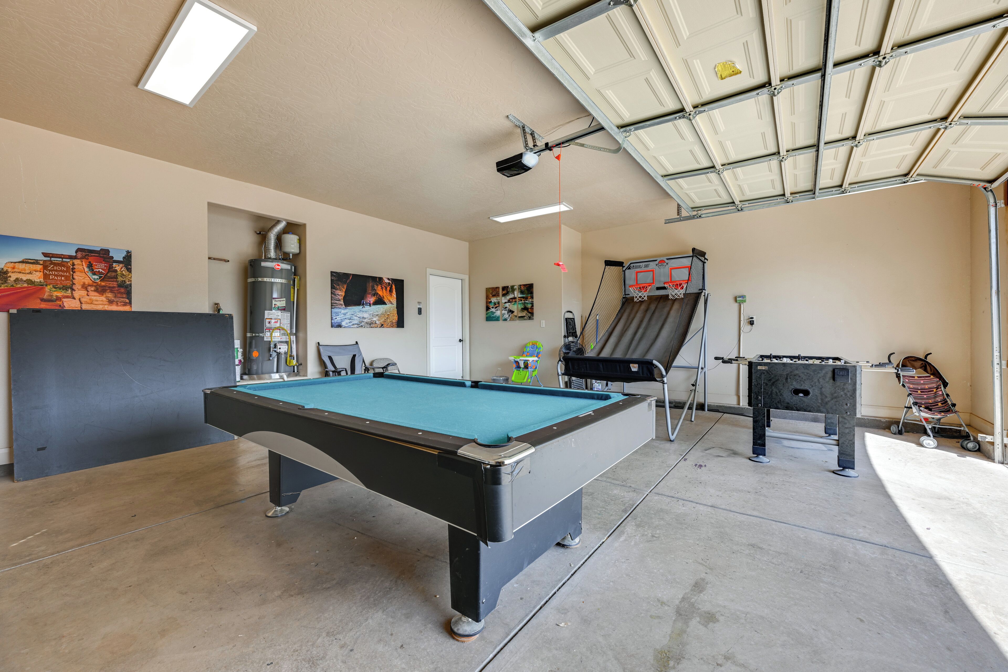 A pool table in a room with a basketball hoop, a foosball table, and a painting on the wall.