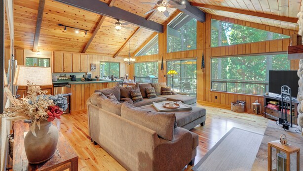 A spacious living area with a large sectional sofa, wooden flooring, and large windows offering views of the outdoors. - Quiet Cabin w/ Deck: Near Lake Almanor! (Westwood)