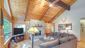 A cozy living room with a stone fireplace, wooden ceiling beams, a large gray sofa, and a flat-screen TV. - Quiet Cabin w/ Deck: Near Lake Almanor! (Westwood)