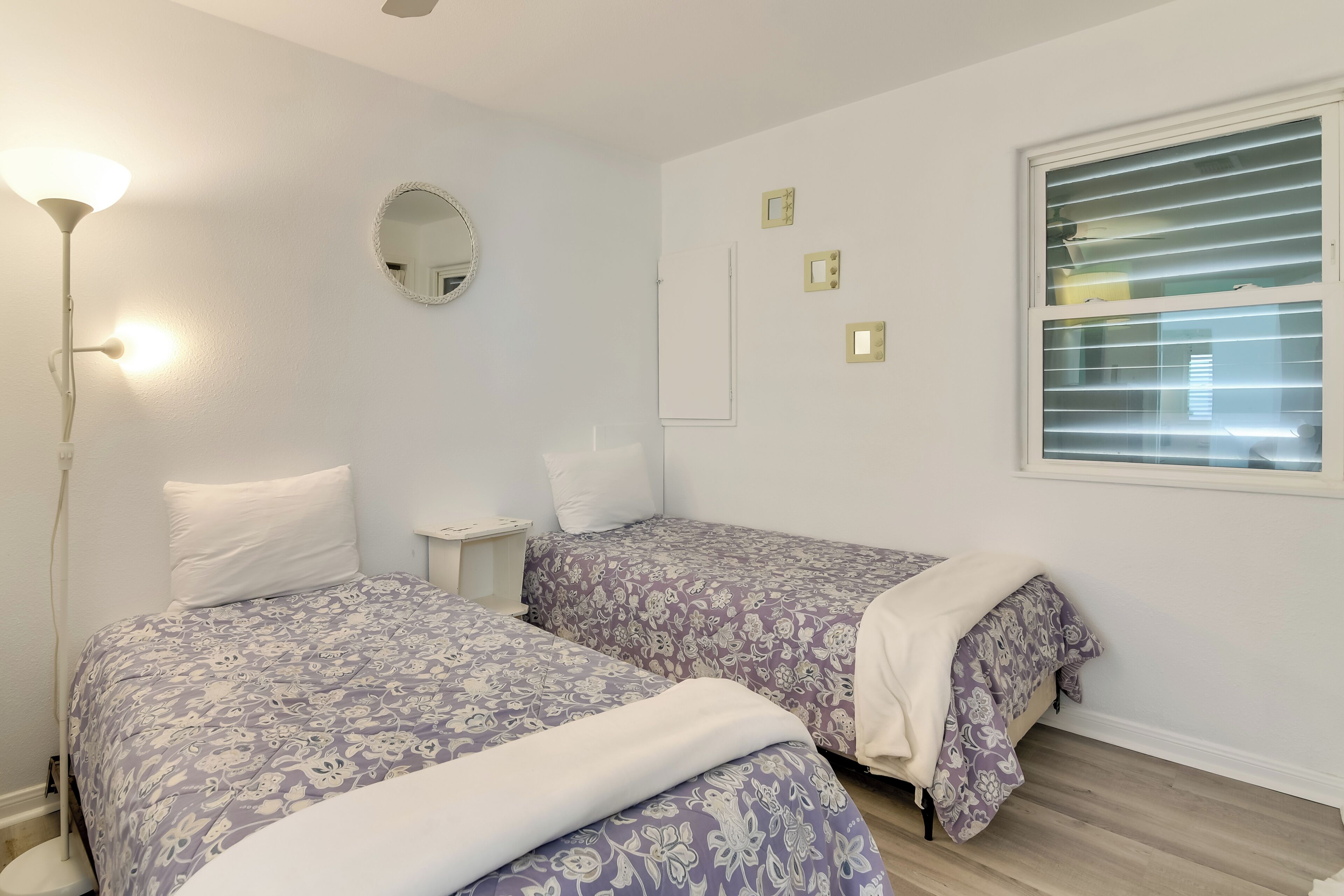 A bedroom with two beds, a bedside table, a lamp, a mirror, and a window with blinds.