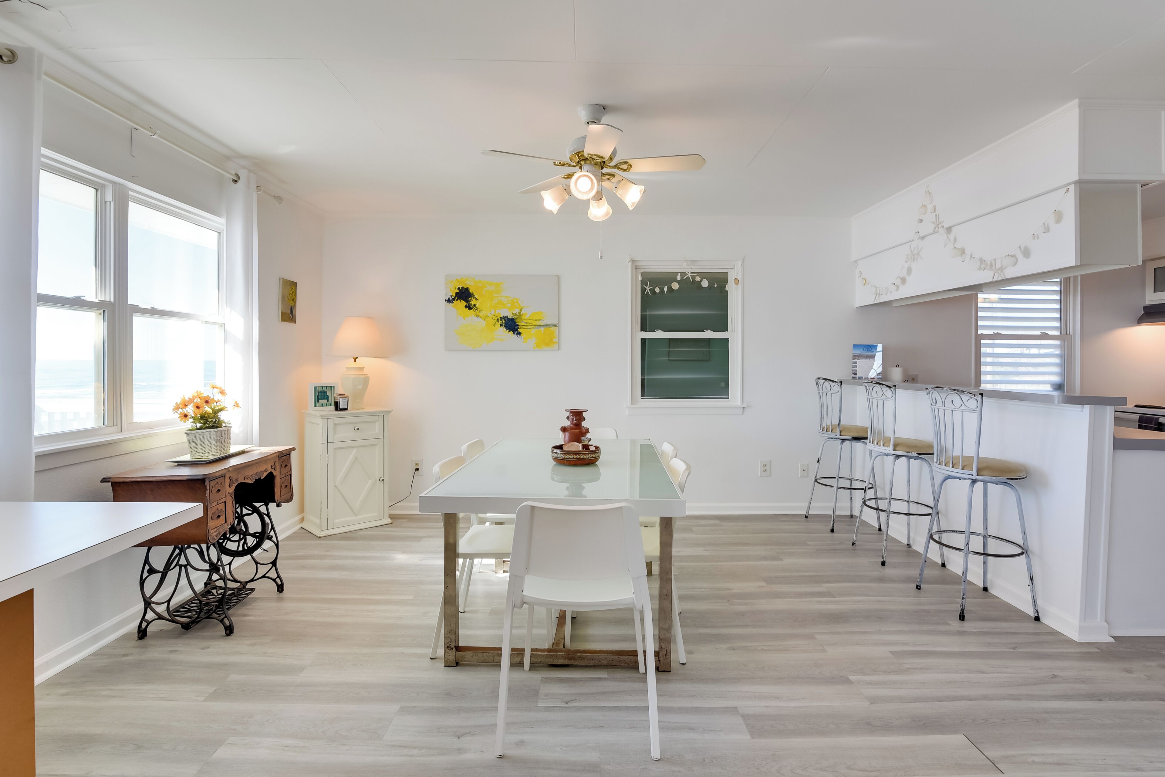 A modern kitchen with a dining table, white chairs, a ceiling fan, and a window with curtains.