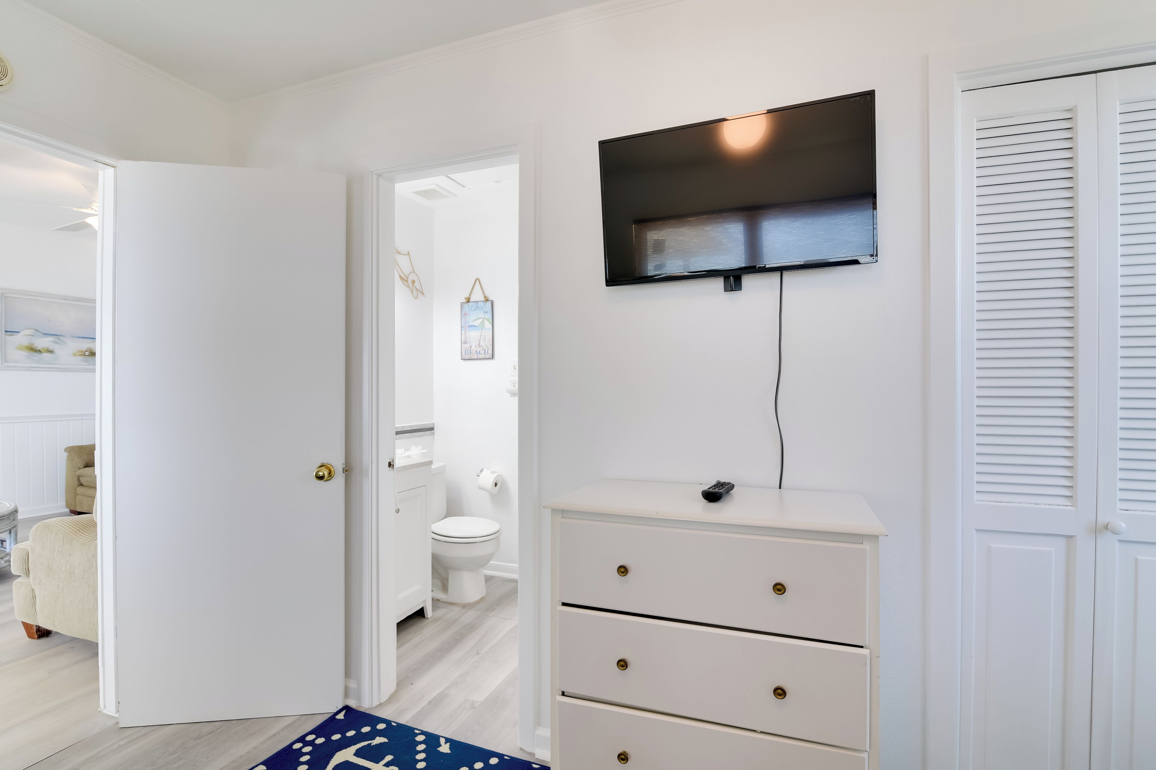 A bedroom with a bed, a dresser, a TV mounted on the wall, and a door leading to another room.