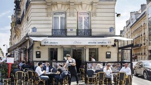 Un café en plein air avec des tables à l’extérieur, un serveur qui s’occupe des clients et une pancarte indiquant « service assuré 7 jours sur 7 ».