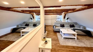 Un ampio loft con travi a vista in legno, due divani grigi, tavolini bianchi e un piccolo tavolino laterale con oggetti decorativi.