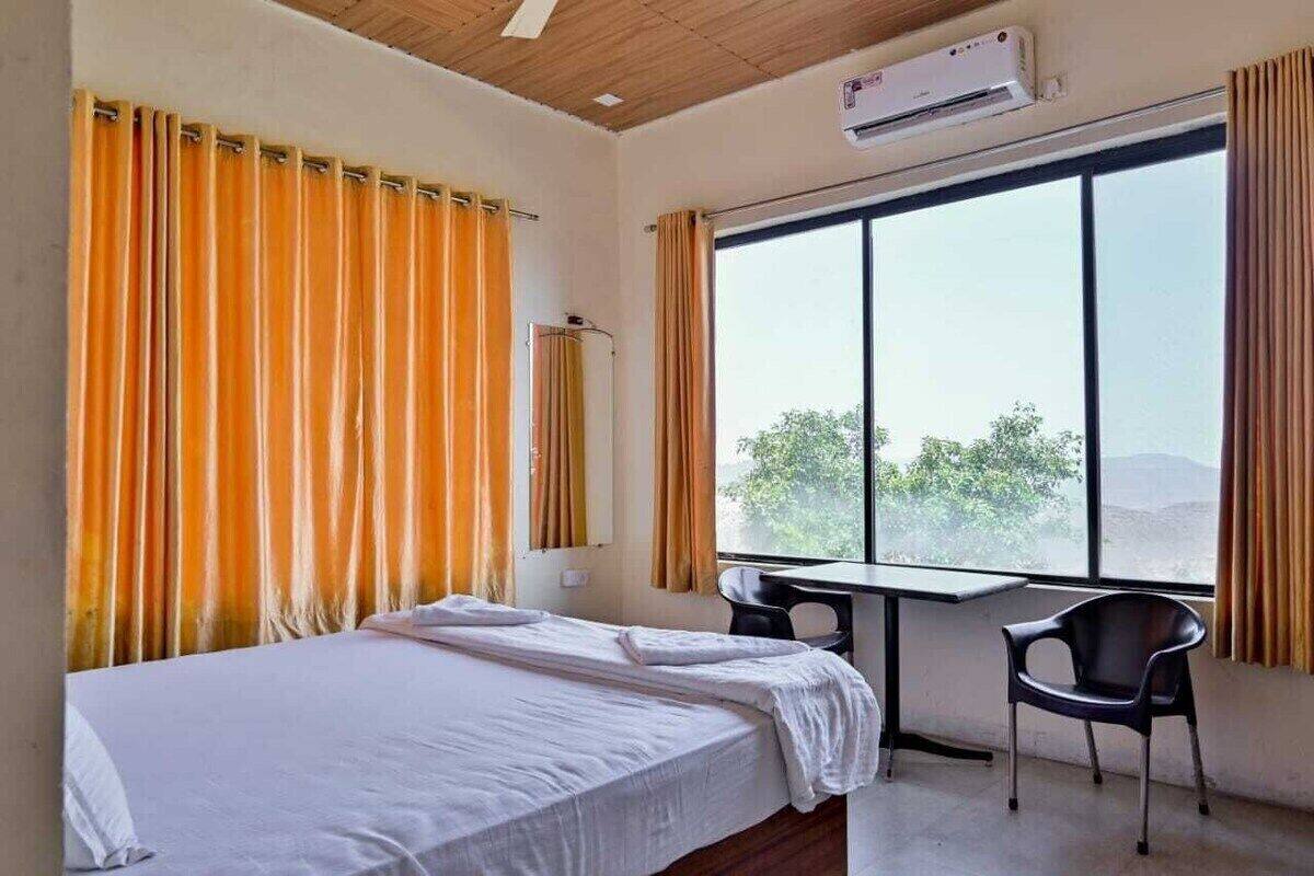 Quarto de hotel com uma cama, uma escrivaninha e uma cadeira. Há uma janela com cortinas, e um ar condicionado fixado na parede.