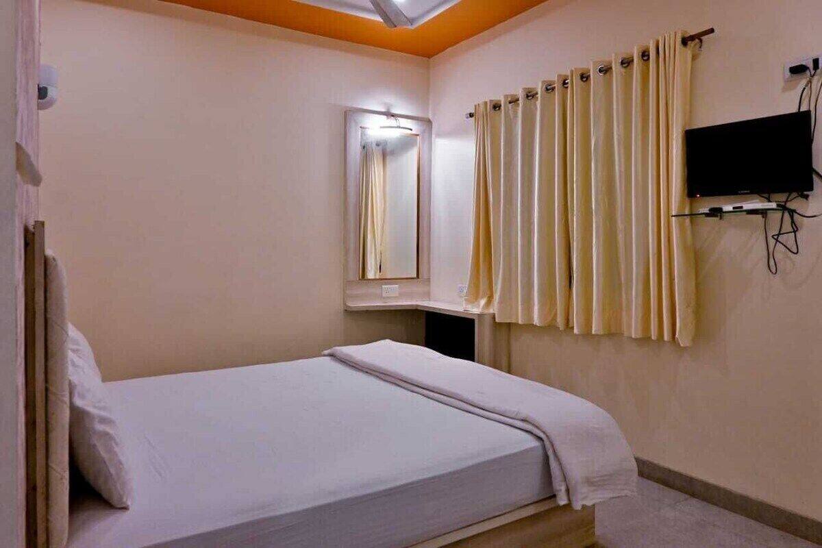 Uma cama de solteiro com colchão e almofadas brancos, uma TV fixada na parede, um espelho e uma cortina.