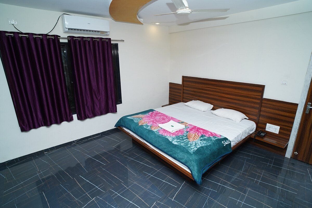 Quarto com cama, cortinas roxas, ventoinha no teto e piso ladrilhado.