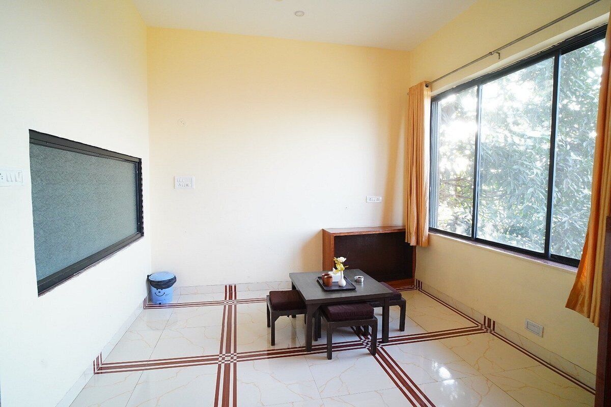 A small room with a tiled floor, a table with chairs, a window with curtains, and a wall-mounted screen.