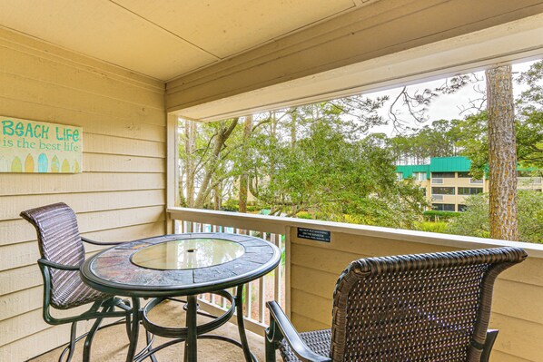 A covered outdoor area with a round table and two wicker chairs, a sign on the wall, and a view of trees and buildings. - Beach & Pool Access: Tranquil Hilton Head Getaway (Hilton Head Island)