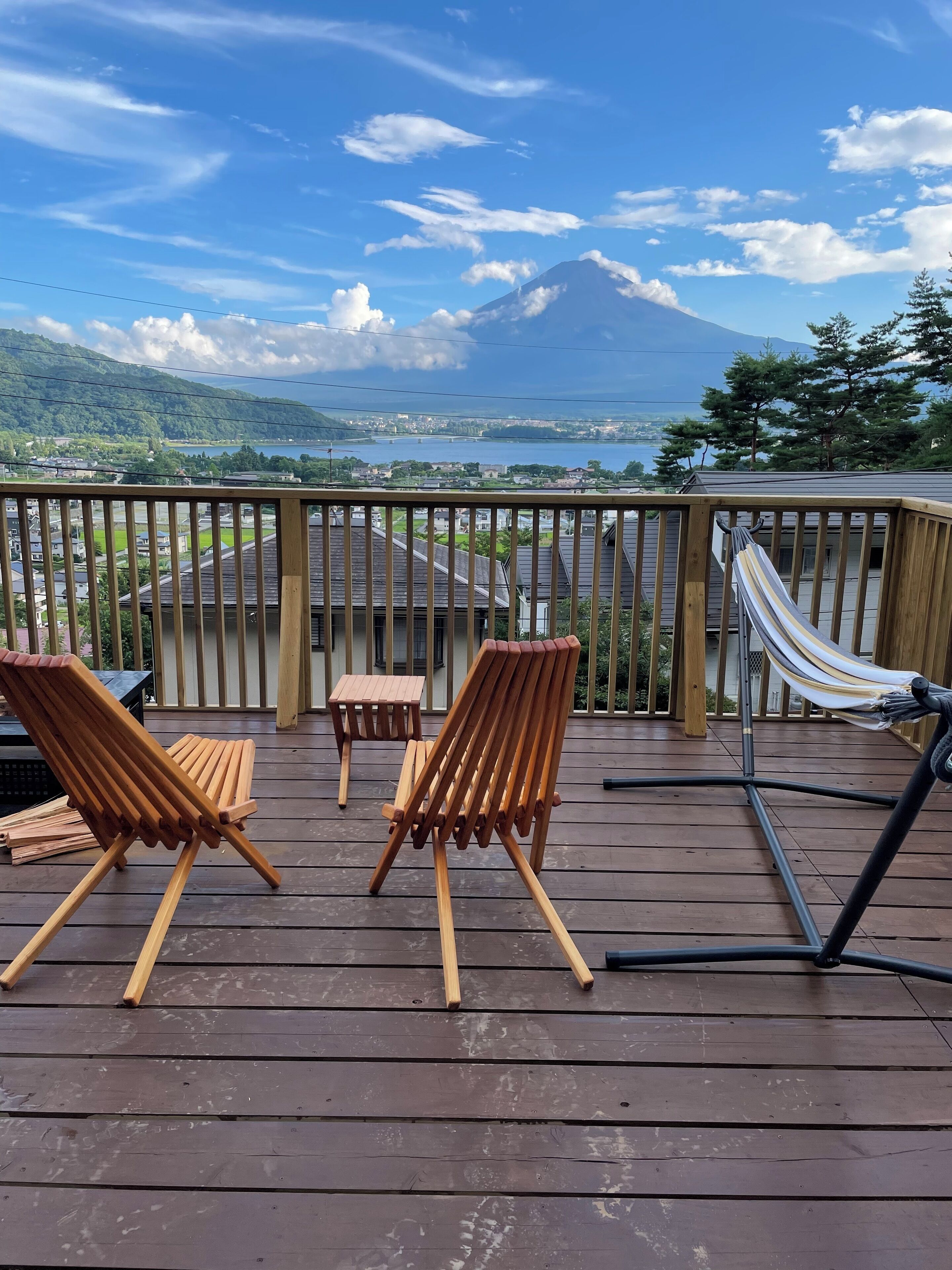 Eine Holzterrasse mit zwei Holzstühlen, einem kleinen Tisch, einer Hängematte und Blick auf einen Berg und See.