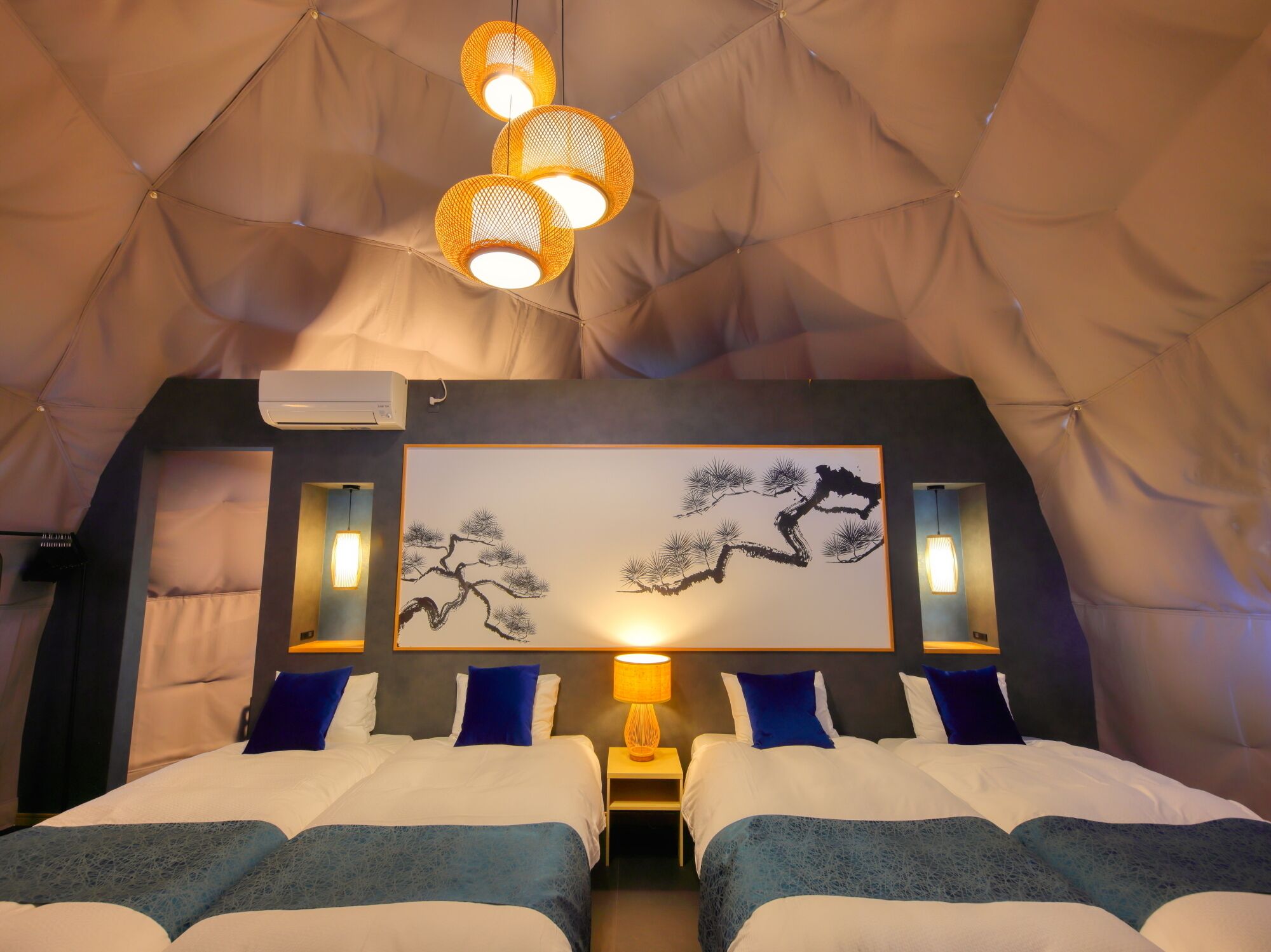Une chambre aménagée comme une tente, avec deux lits, un climatiseur fixé au mur et un tableau représentant des arbres.
