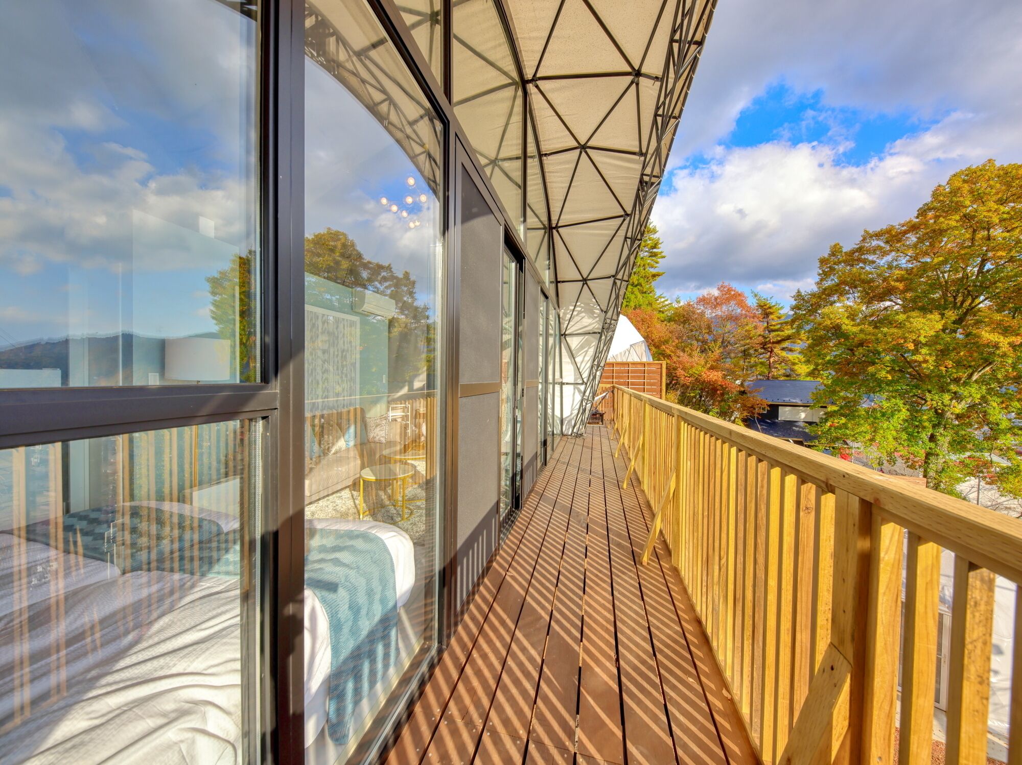 A wooden deck with glass railings, a covered area with a geometric roof, and a view of trees and buildings.