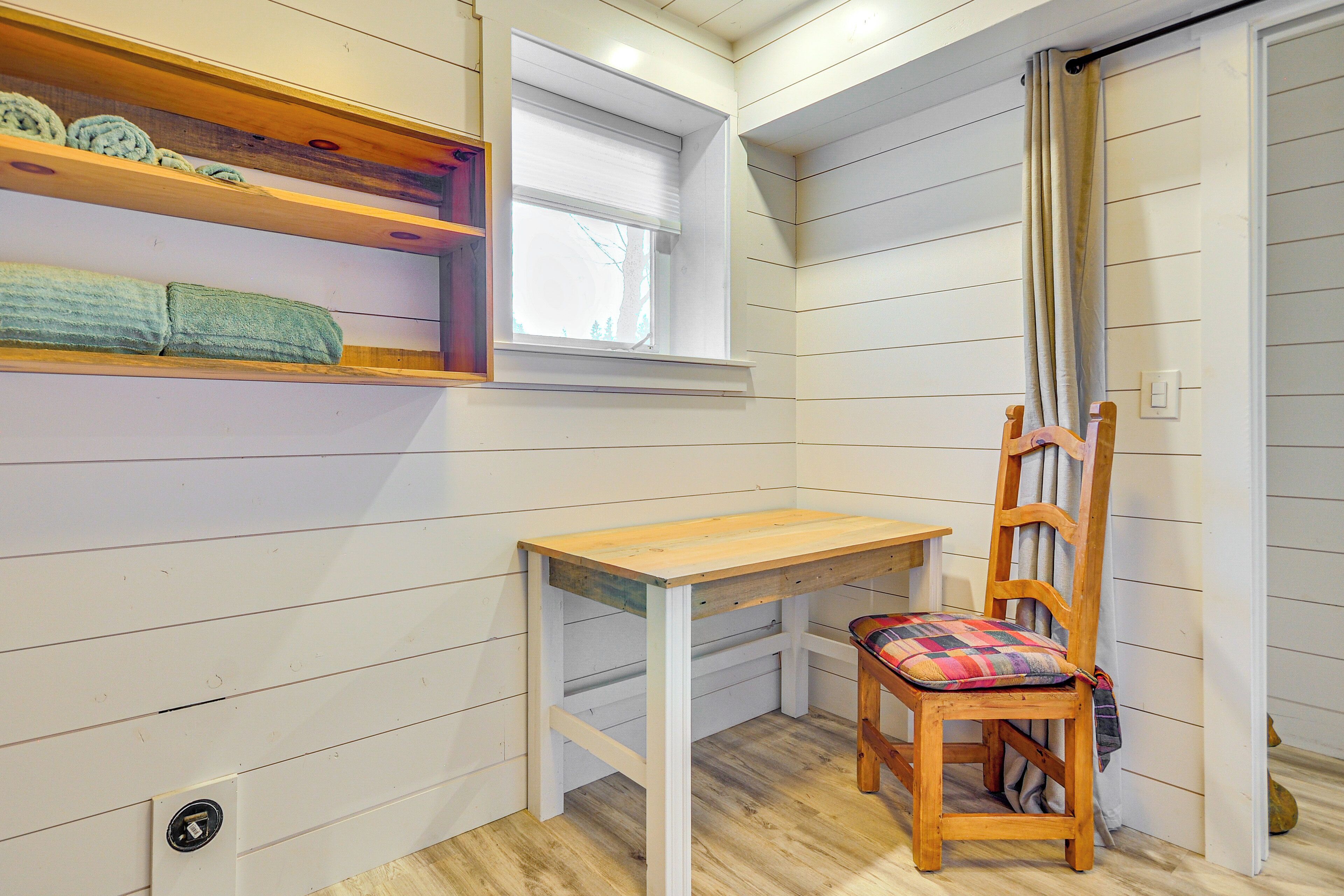 A corner room with a wooden table, a chair, a window, and a shelf with towels.