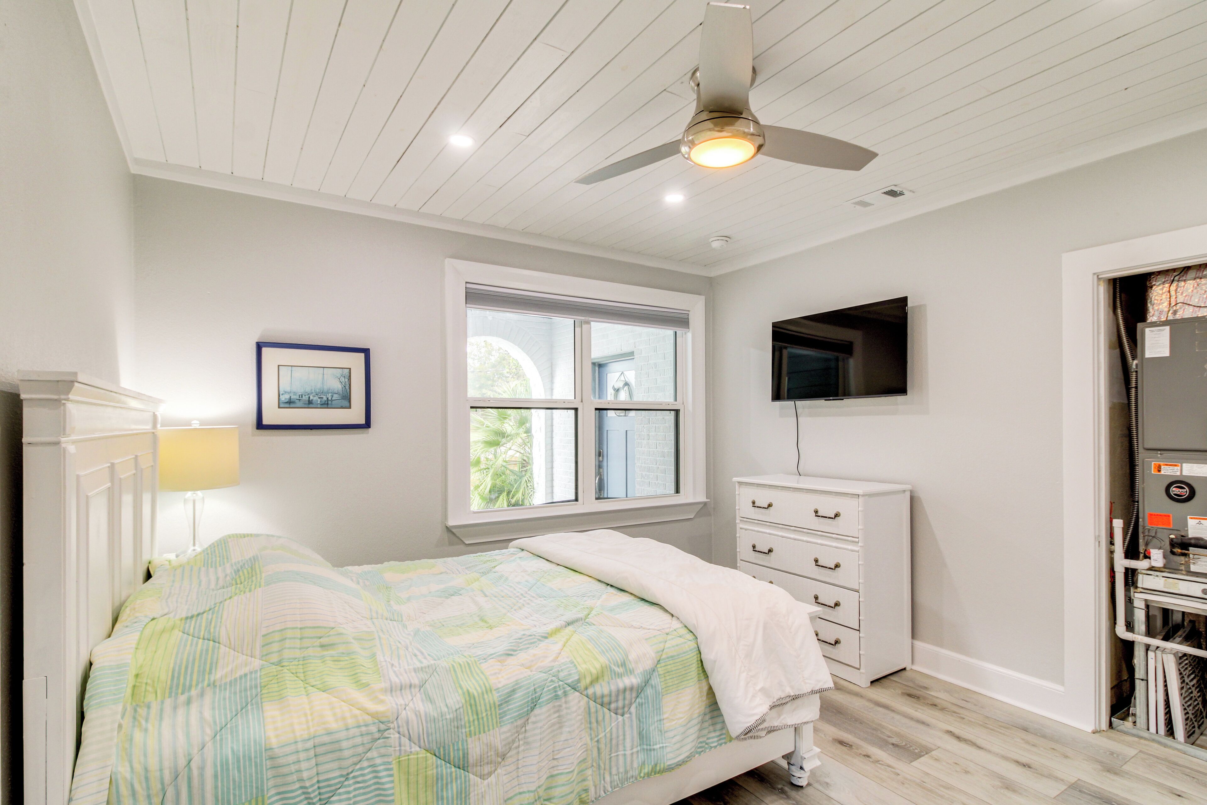 A bedroom with a bed, a ceiling fan, a window, a dresser, and a TV mounted on the wall.