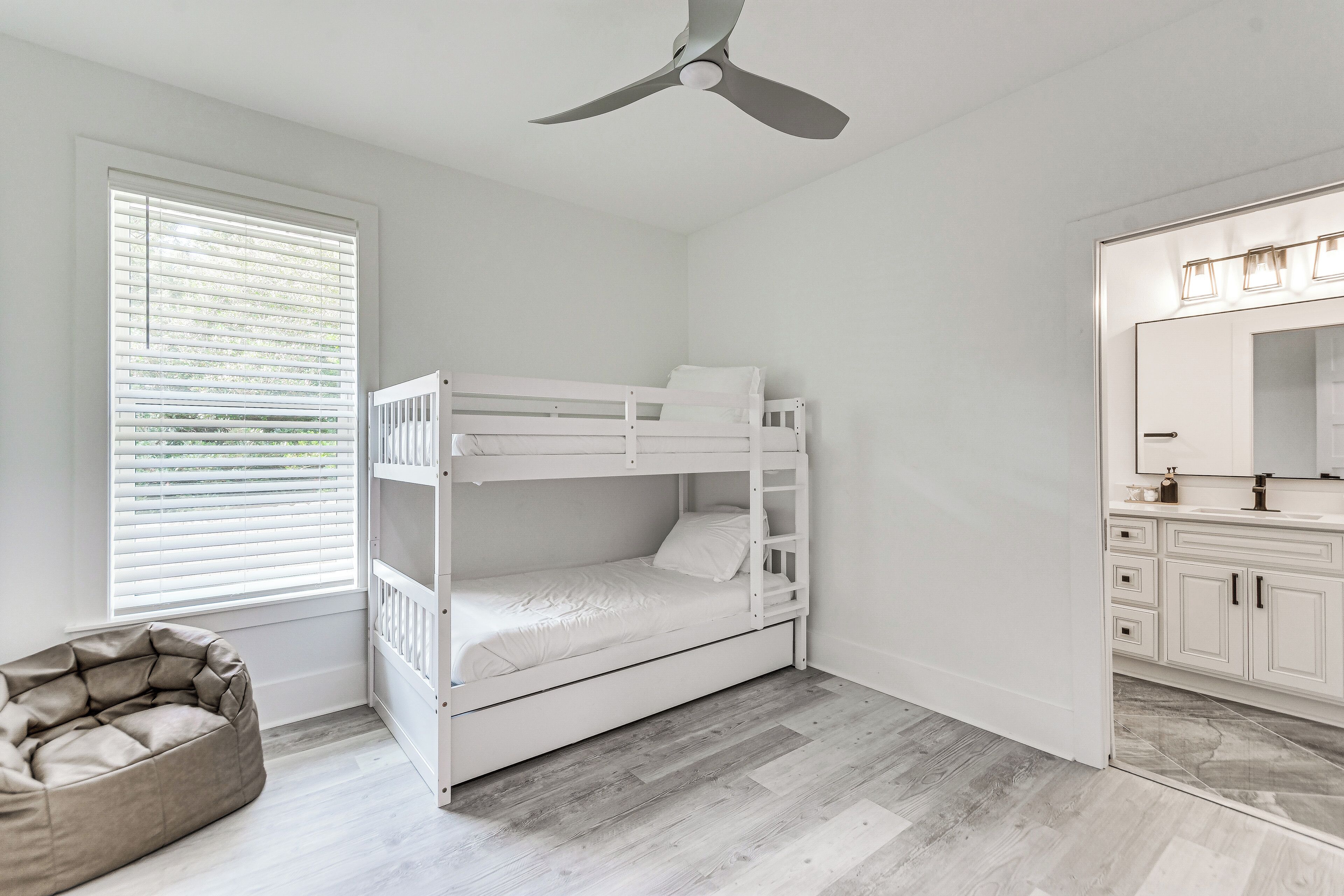 Une chambre avec un lit superposé, un ventilateur de plafond, une fenêtre avec des stores, un fauteuil en forme de poire et une salle de bain avec un meuble-lavabo et un miroir.