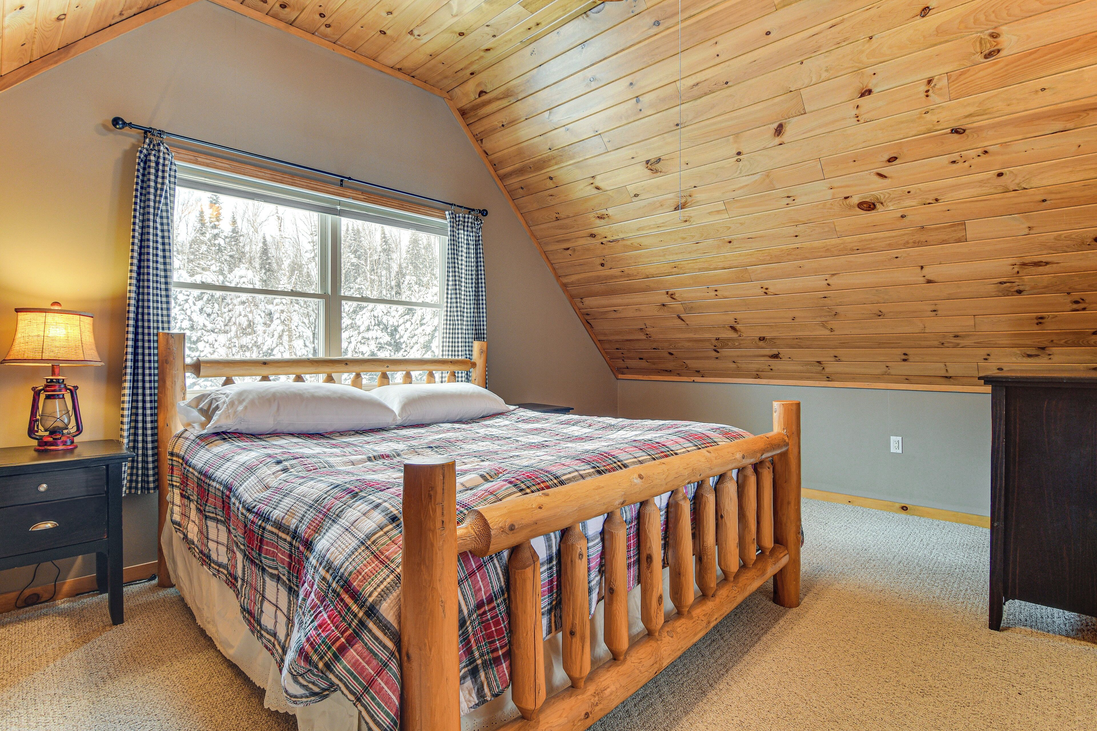 Une chambre avec un plafond en bois, un lit recouvert d’une couverture à carreaux, une table de chevet avec une lampe et une fenêtre donnant sur des arbres.