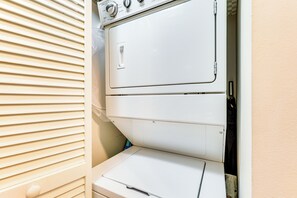 A white stacked washer and dryer unit in a laundry room with a beige door and blinds.