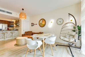 A modern living room with a dining area, featuring a hanging chair, white chairs, a round table, and a kitchen in the background.