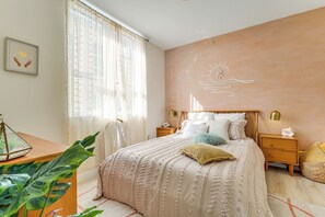 A bedroom with a large bed, a nightstand, a bedside table, and a window with sheer curtains.