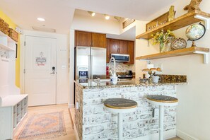 A kitchen with a white brick backsplash, wooden cabinets, a stainless steel refrigerator, a microwave, and a coffee maker on the counter. There are two stools at the island and a globe on the shelf above.