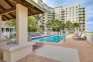 A pool area with lounge chairs and a covered seating area, surrounded by palm trees and a multi-story building with balconies.