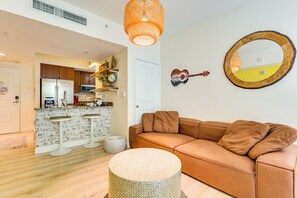 A modern kitchen with a refrigerator, microwave, and wooden cabinets. There is a brown leather sofa and a round woven ottoman in the living area.
