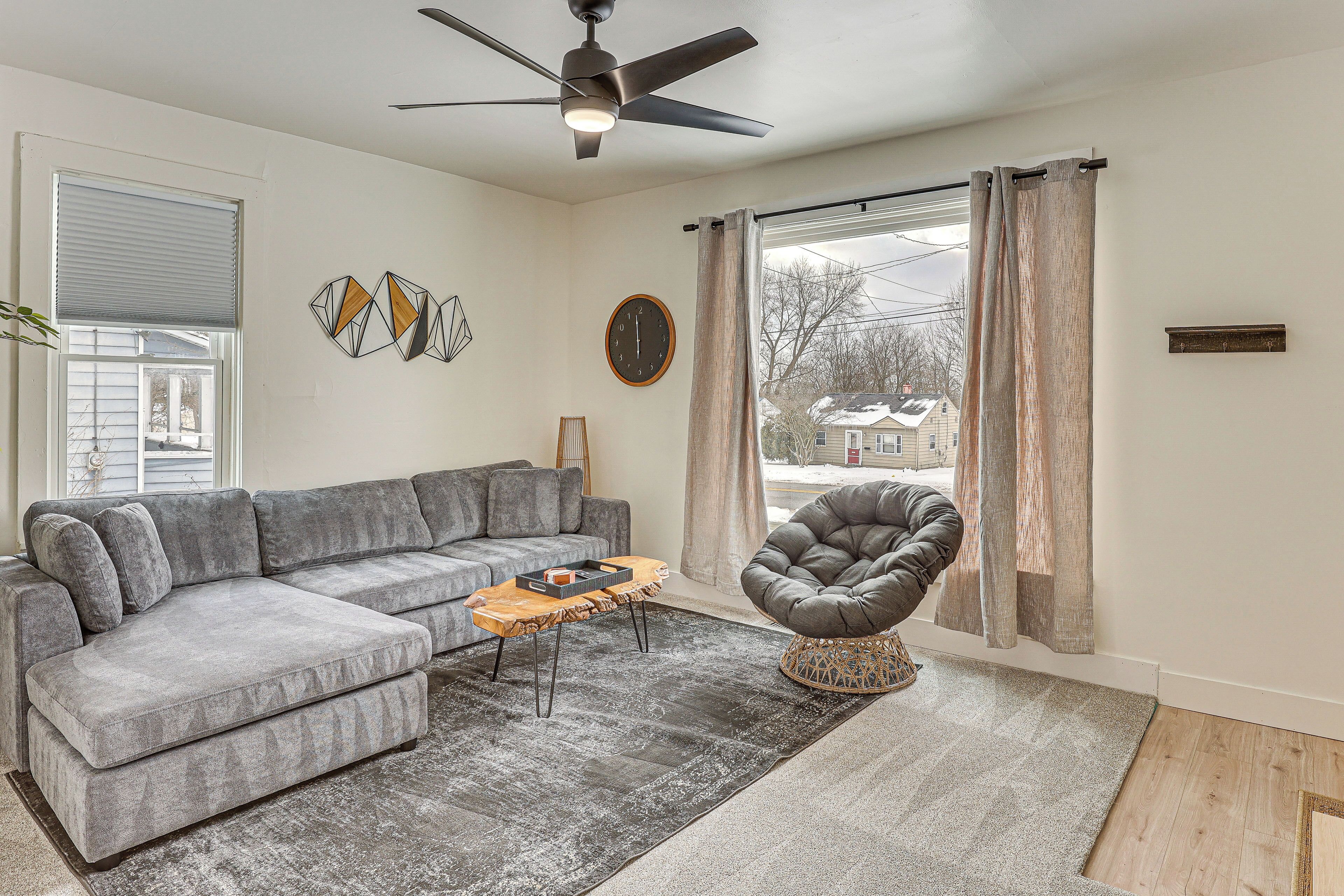 Un salon avec un canapé d’angle gris, une table basse en bois, un fauteuil gris, un ventilateur de plafond et une grande fenêtre avec des rideaux.