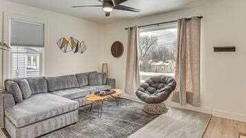 Un salon avec un canapé d’angle gris, une table basse en bois, un fauteuil gris, un ventilateur de plafond et une grande fenêtre avec des rideaux.