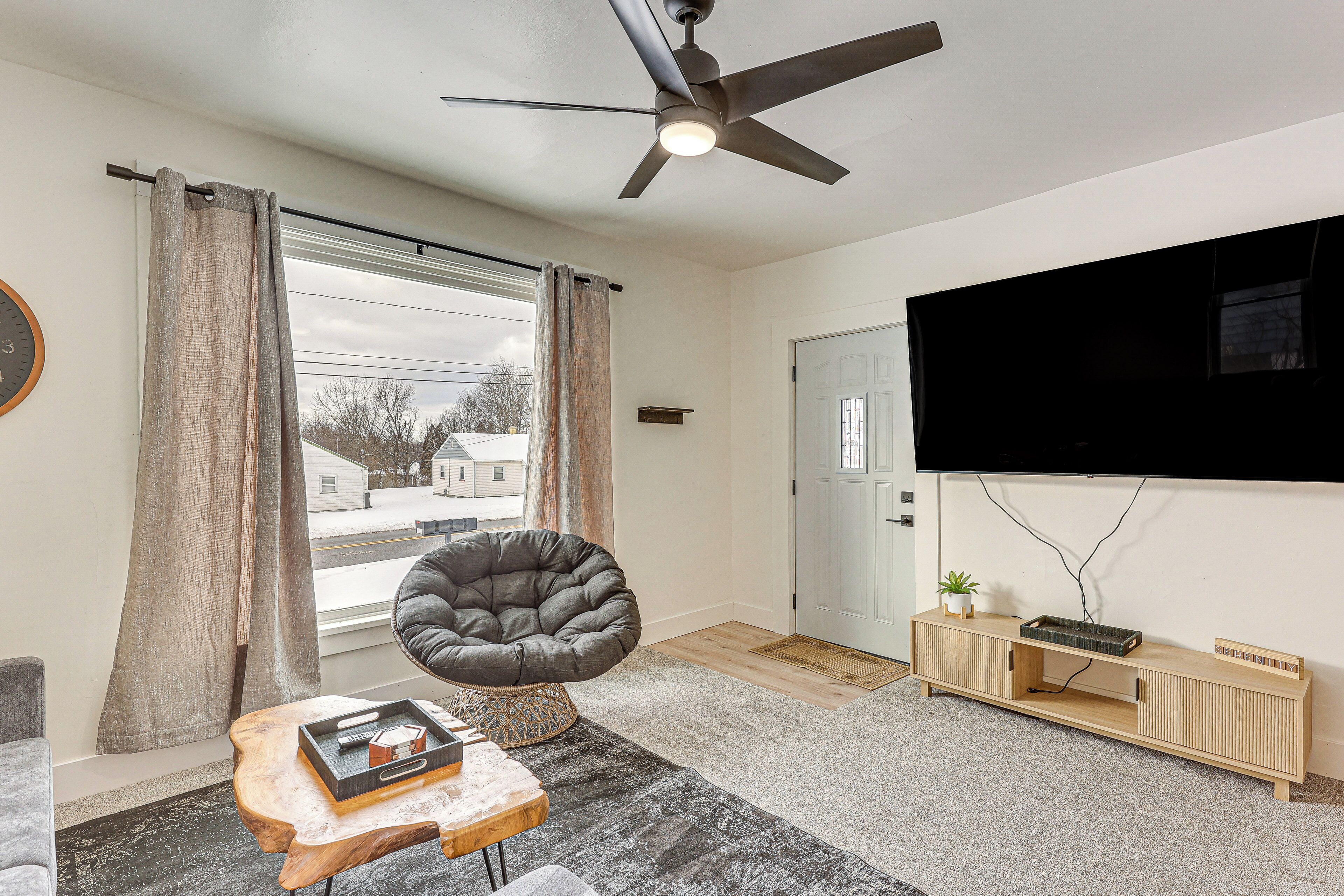 Un salon moderne doté d’un ventilateur de plafond, d’une grande fenêtre avec des rideaux, d’un fauteuil confortable, d’une table basse en bois et d’un téléviseur fixé au mur.