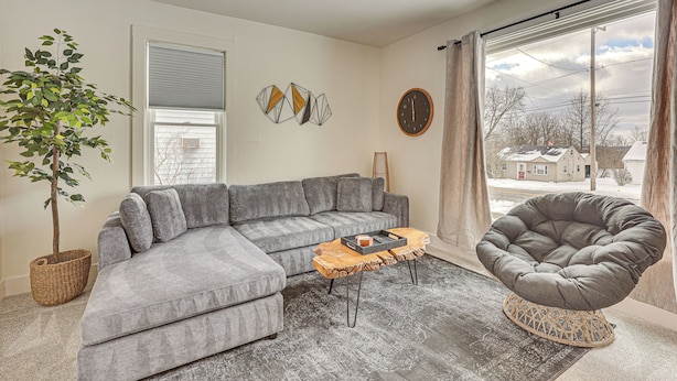 Un salon avec un canapé d’angle gris, un fauteuil en forme de poire, une table basse en bois, une plante en pot et un ventilateur de plafond.