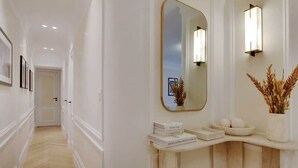 Un couloir avec un mur blanc, un grand miroir et une coiffeuse ornée d’un vase contenant des plantes séchées.
