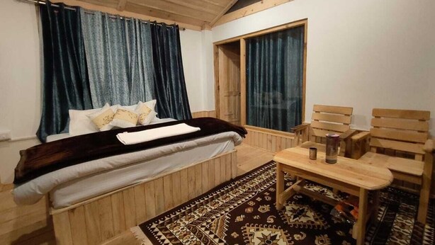 Une chambre à coucher avec un lit, une tête de lit en bois, une petite table avec des chaises, un tapis à motifs et une fenêtre avec des rideaux.