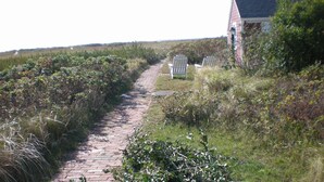 A narrow brick pathway leading to a house with two white Adirondack chairs. - Unique Coastal Retreat in Exclusive Condo Complex (2392) (Truro)