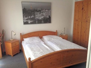 A bedroom with a wooden bed, two bedside tables, a nightstand, and a cityscape painting on the wall.