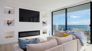A modern living room with a large flat-screen TV, a sofa with cushions, and a view of the sea through large windows.