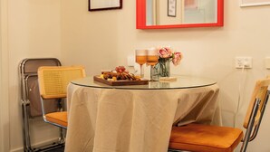 Una mesa redonda con mantel beige, dos sillas, una bandeja con aperitivos, dos vasos y un jarrón con flores.