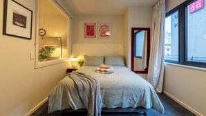 Un dormitorio ordenado con cama, mesita de noche, lámpara y vista a la ciudad a través de una ventana.