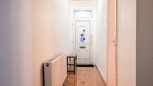 En smal hall med en vit dörr som har ett fönster med målat glas och blommönster. En liten trähylla är placerad mot väggen nära ett element.