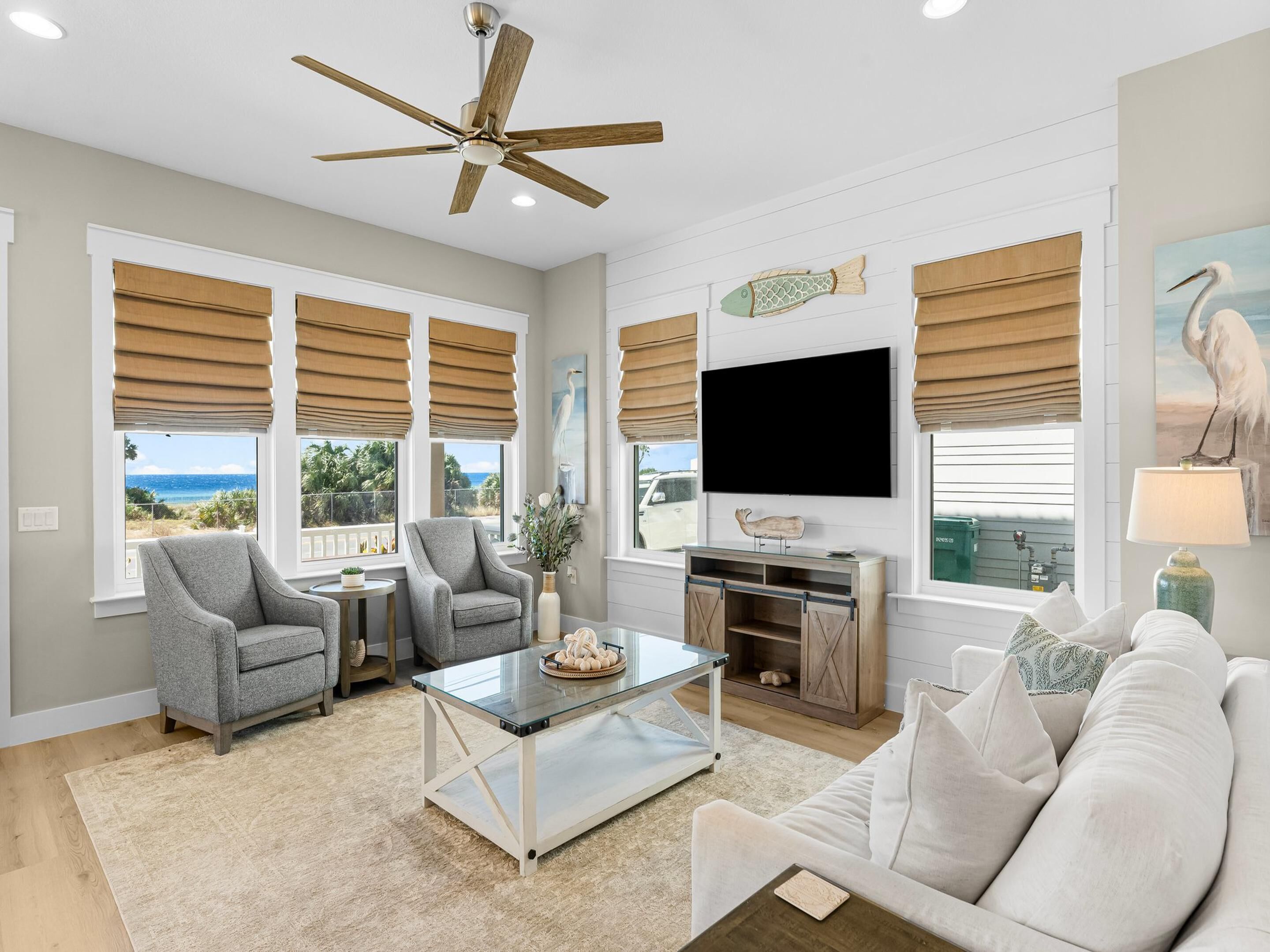 A spacious living room with a ceiling fan, large windows with blinds, a flat-screen TV, and a comfortable seating area.