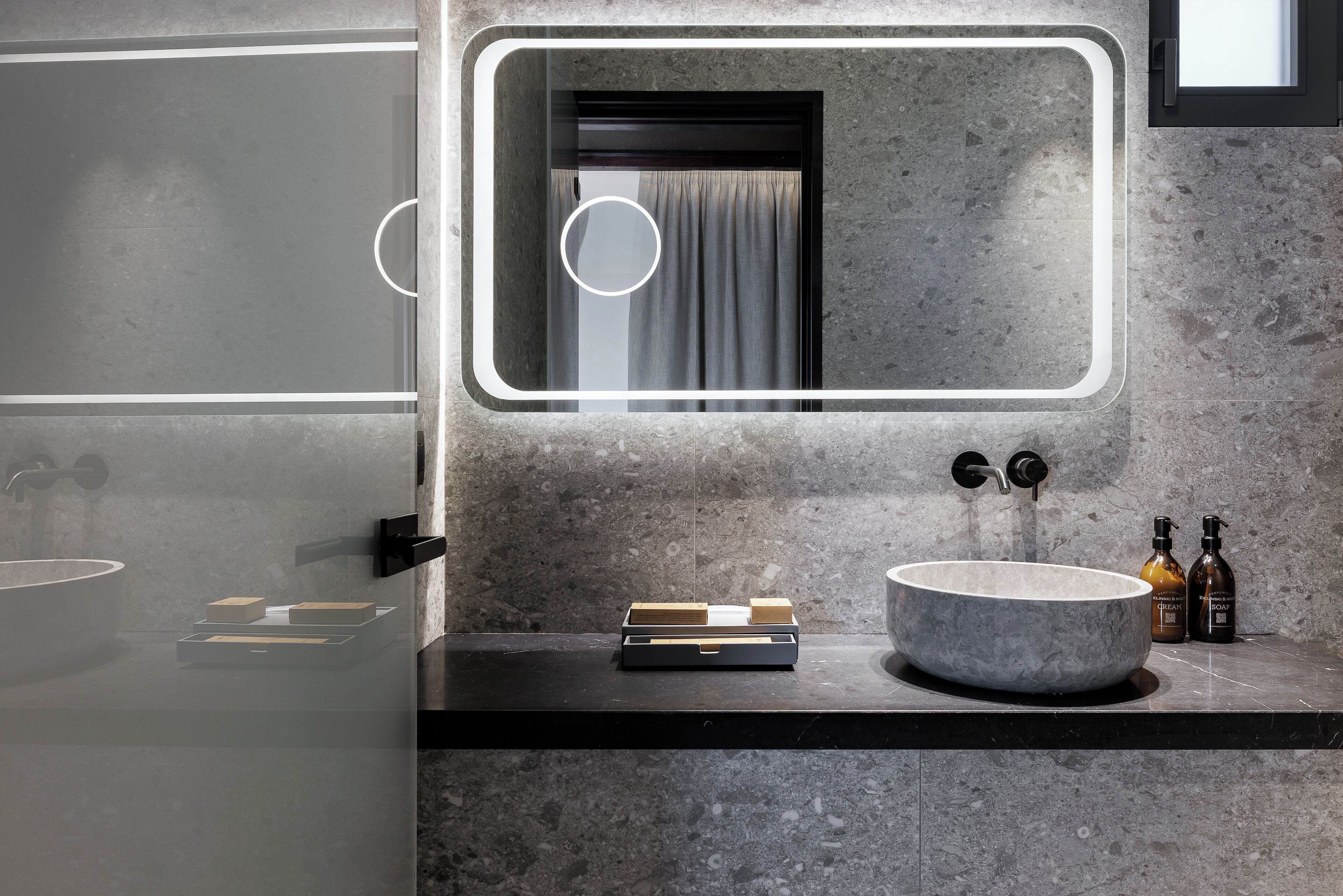 A modern bathroom with a large round sink, a mirror with a circular light, and a small window with curtains.