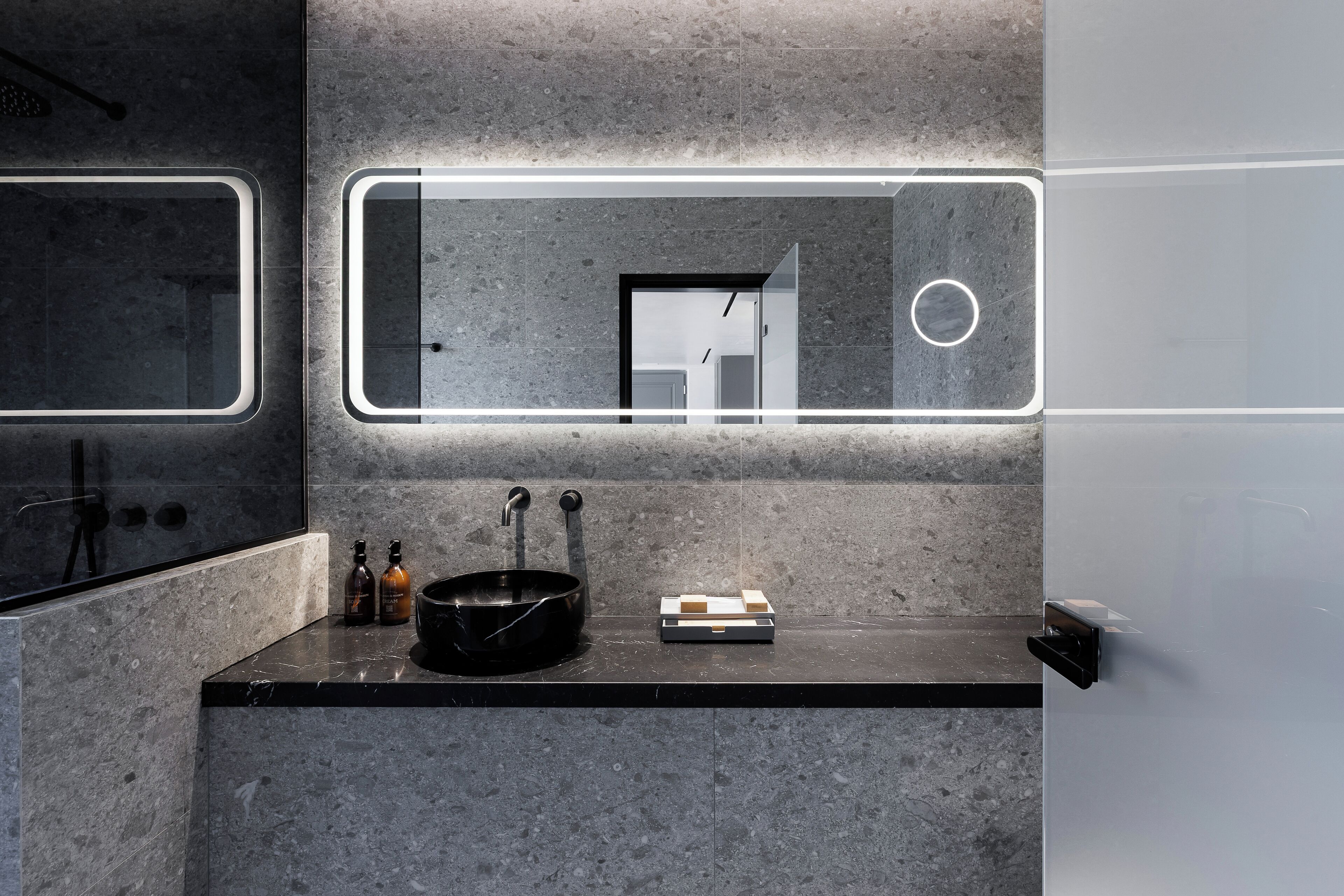 A modern bathroom with a large mirror, a black sink, and two bottles on the counter.