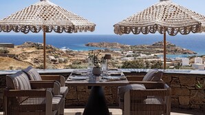 Mesa lista para comer con dos grandes sombrillas tejidas, sillas de mimbre y vista al mar y al paisaje.