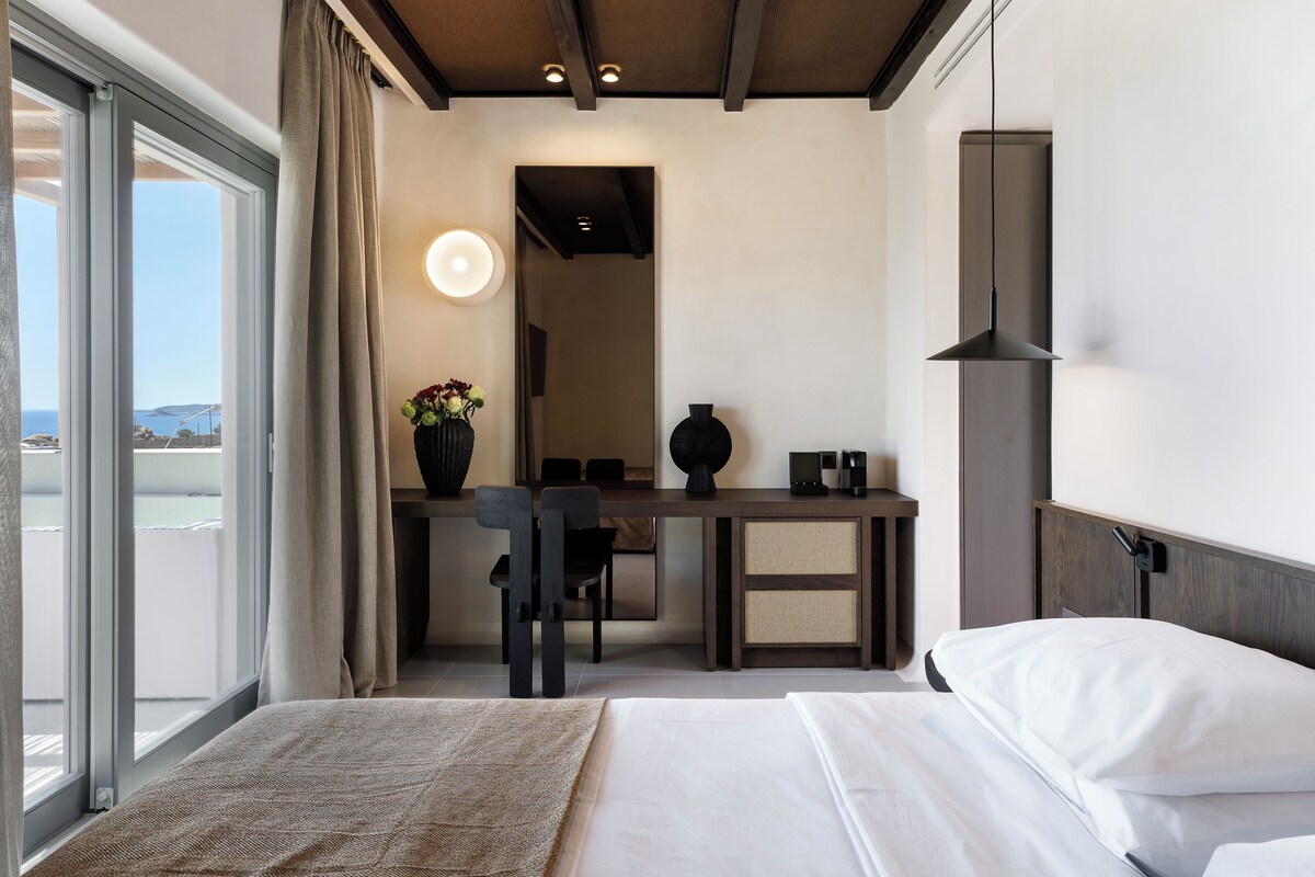 Een hotelkamer met een bed, een bureau, een stoel, een vaas met bloemen en uitzicht op zee.