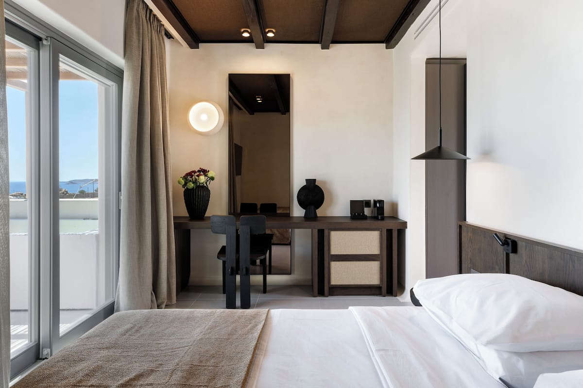 Een hotelkamer met een bed, een bureau, een stoel, een vaas met bloemen en uitzicht op zee.