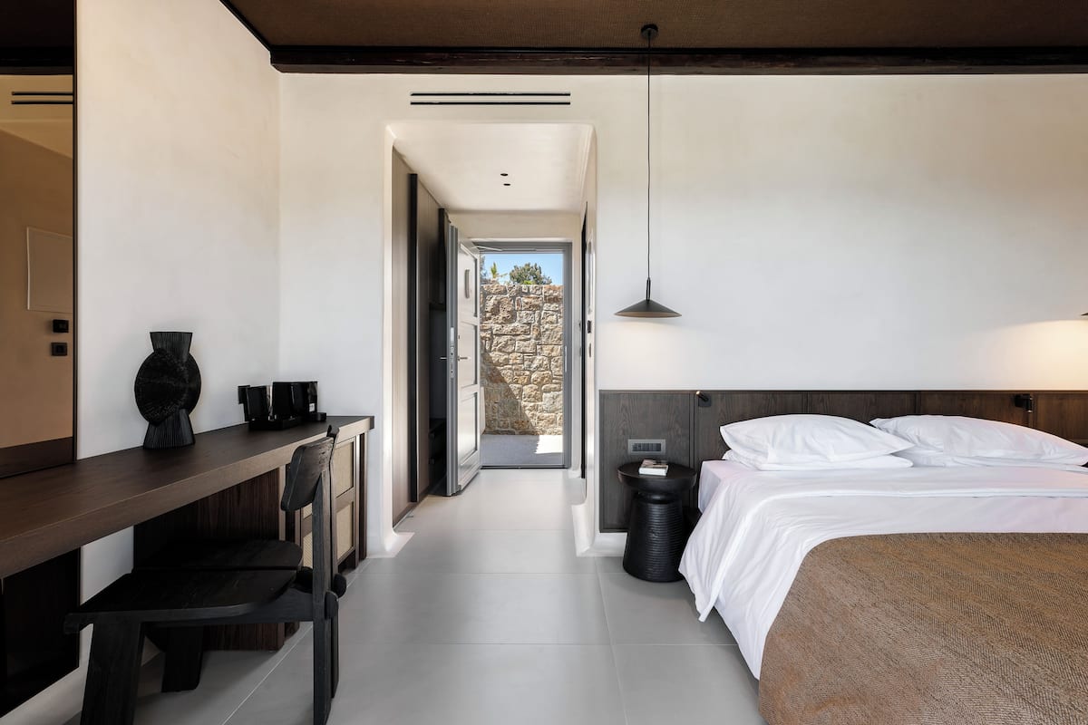 Een moderne hotelkamer met een houten bureau, een bed met witte lakens en uitzicht op een stenen muur door een open deur.