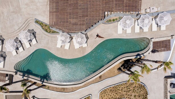 Vista aérea de un resort con piscina curvo, sillones de descanso y palmeras.