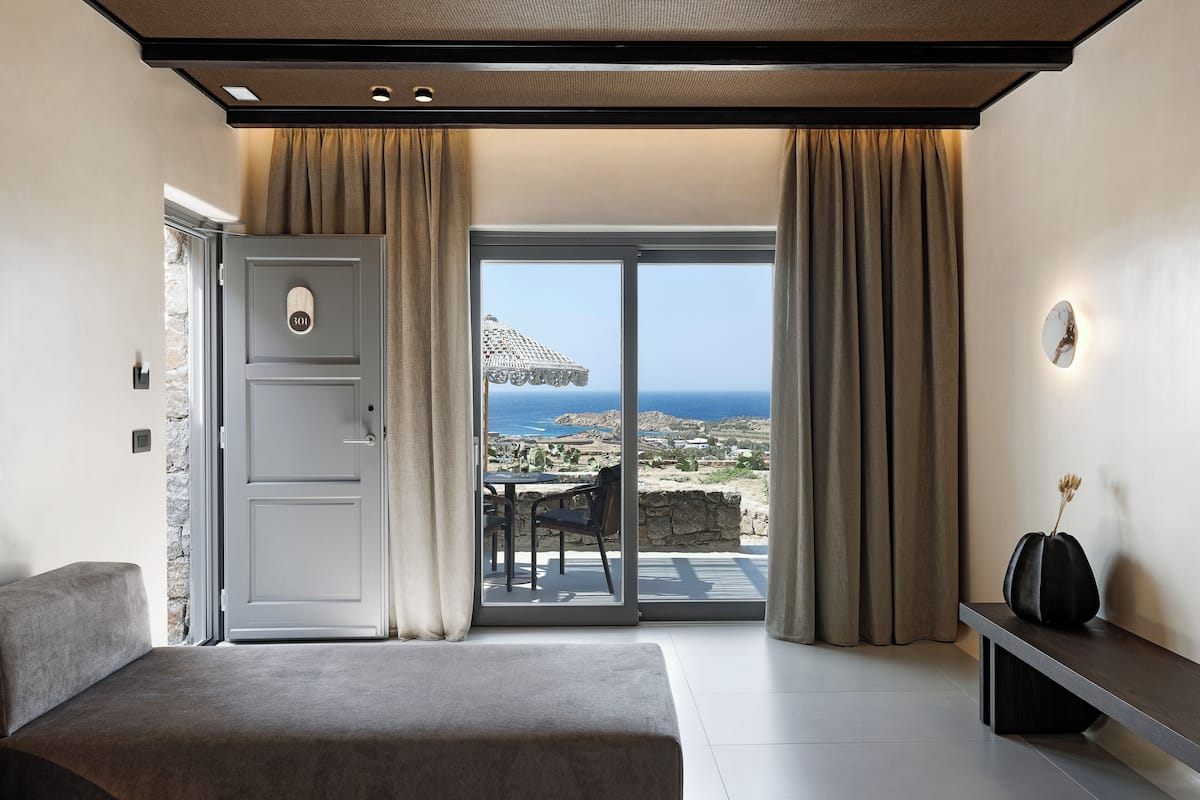 Een slaapkamer met een bed, een bankje, een tafel met stoelen en uitzicht op de zee door een glazen deur.